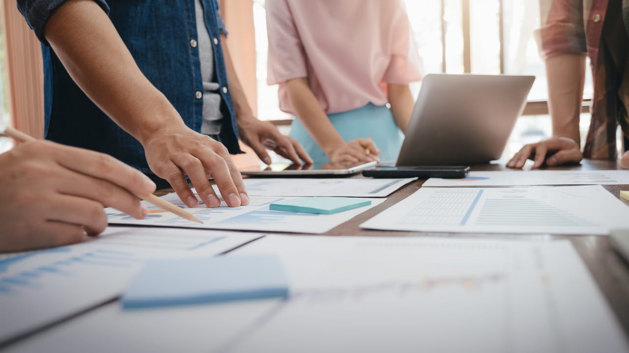 Personer som står runt ett bord med papper, pennor och datorer på