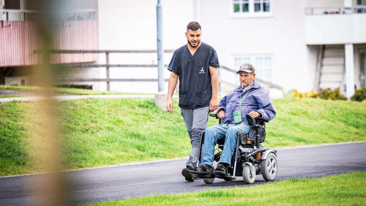 Äldre herre i permobil ledsagas av vårdbiträde