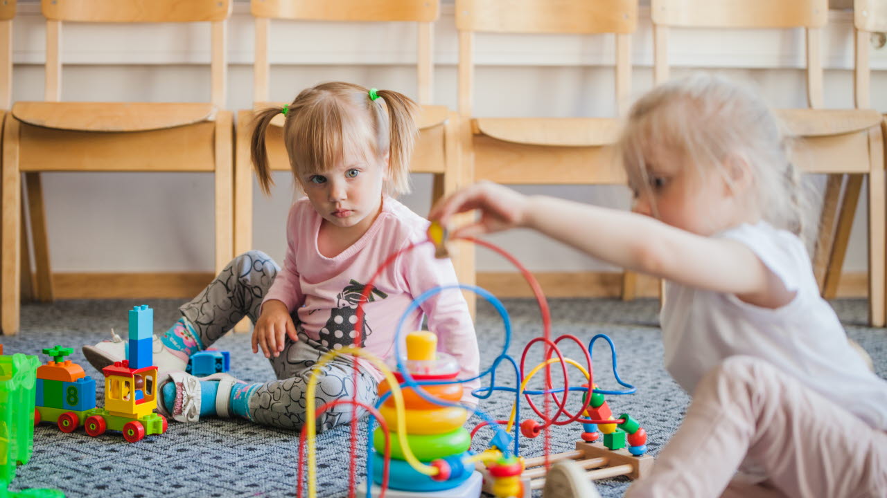 Två små barn sitter på golvet och leker med färgglada leksaker i ett rum med trästolar i bakgrunden.