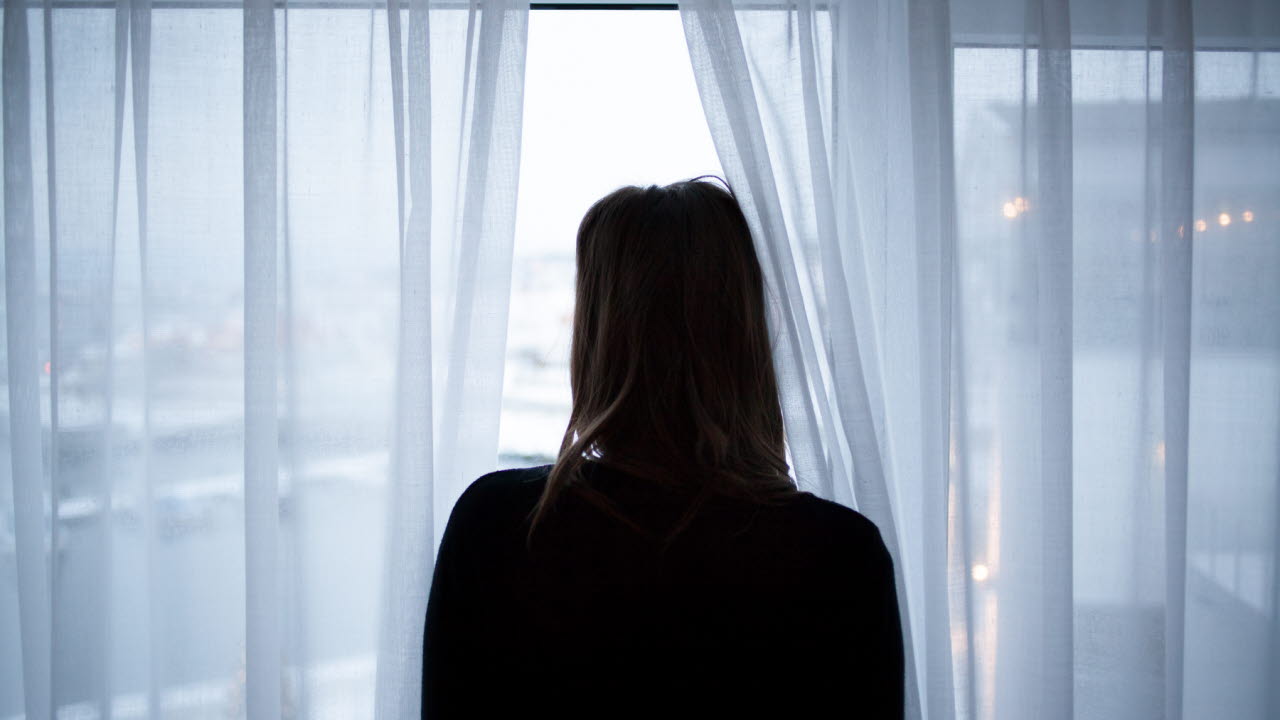 Mörk statyett av en person som tittar ut genom ett fönster med tunna vita gardiner.