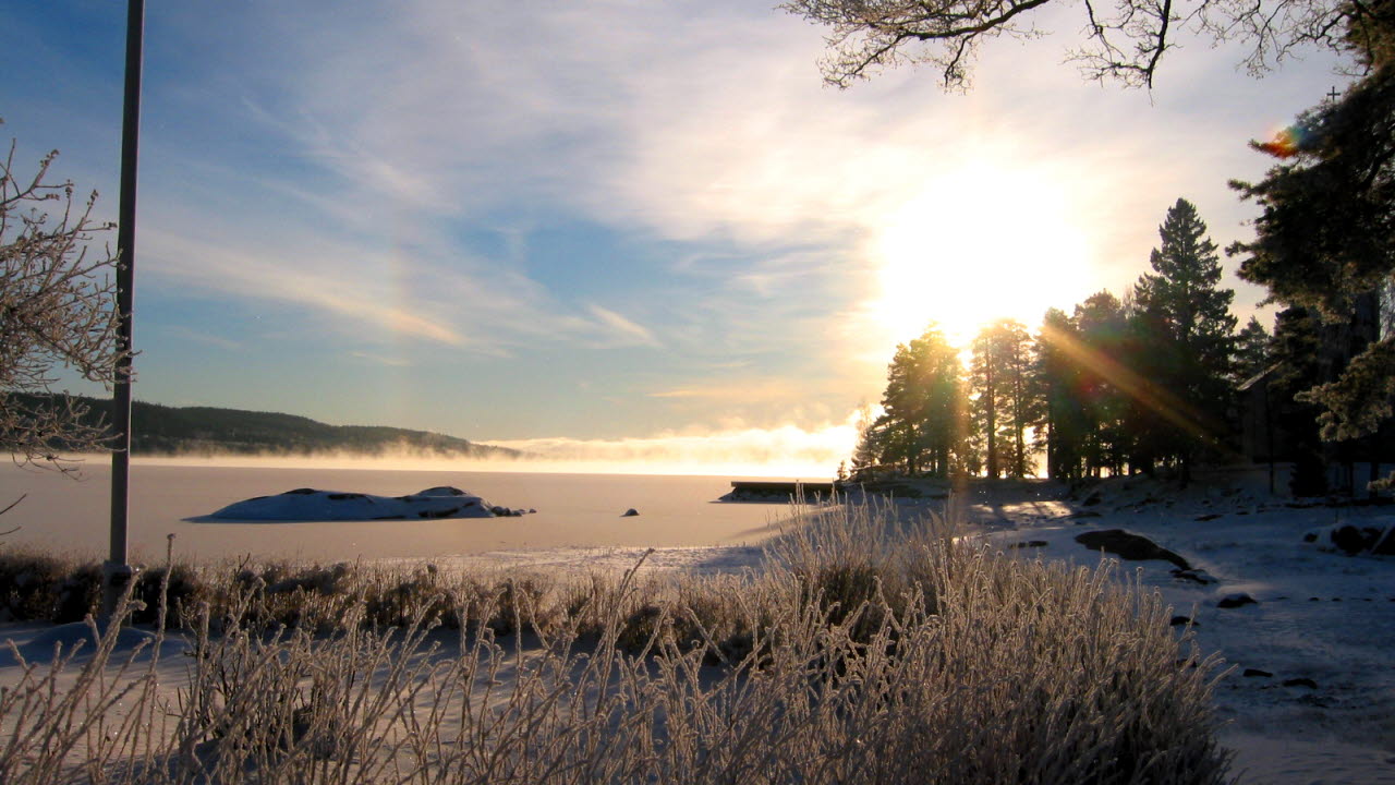 Det är vinter och solen tittar fram bakom barrskogen. Vi står på stranden vid en istäckt sjö.