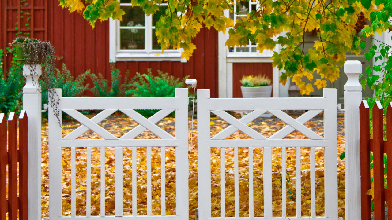 Ett vitt staket som leder till ett rött hus med vita knutar och en trädgård i höstfärger.